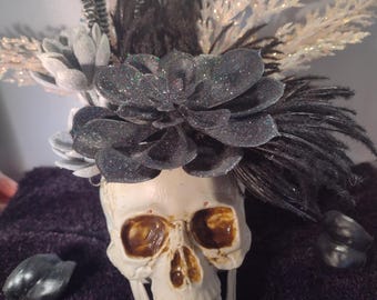 Pode incluir: Uma caveira branca com suculentas de glitter preto e penas brancas e pretas no topo. A caveira está sobre uma superfície preta.
