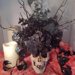 Pode incluir: Uma caveira branca com flores pretas de glitter e galhos com morcegos pretos no topo. A caveira está sentada em um tecido vermelho e branco.