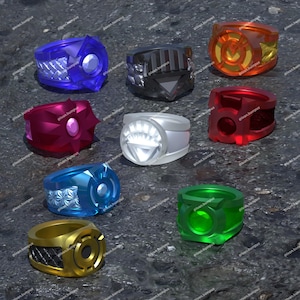 Puede incluir: Una colección de nueve anillos en varios colores, incluyendo azul, negro, naranja, magenta, blanco, rojo, azul claro, verde y dorado. Cada anillo presenta un diseño geométrico distintivo, un emblema central y detalles texturizados.
