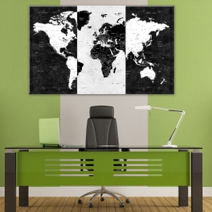 May include: A black and white world map print in three panels hangs on a wall above a glass-top desk with a green base. A black office chair sits in front of the desk. A lamp with a white shade is on the desk.