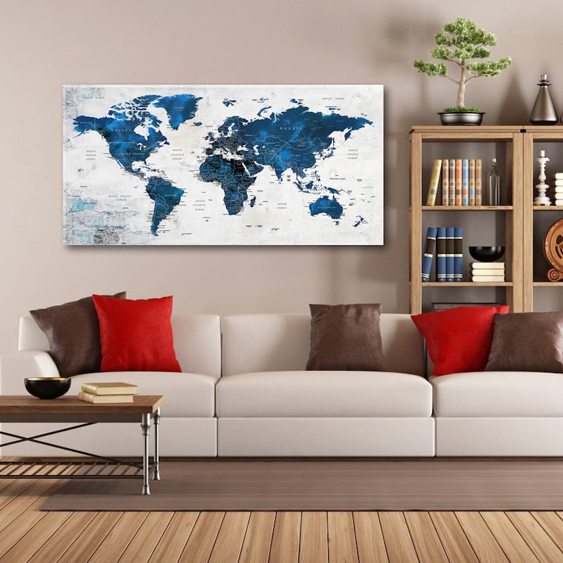May include: A white canvas print of a world map with blue continents and white countries. The map is on a wall above a white couch with red and brown throw pillows. A wooden bookshelf with books is on the right side of the image.