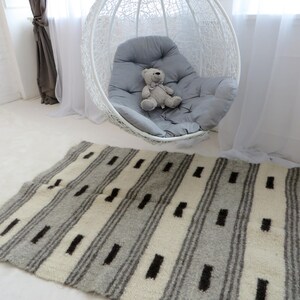 Puede incluir: Una alfombra rayada blanca y gris con detalles negros. La alfombra está hecha de lana y tiene una textura suave y esponjosa. Está colocada sobre un suelo blanco delante de una silla colgante blanca con un cojín gris.