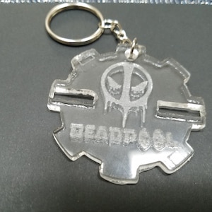 May include: A clear acrylic keychain with a silver metal ring. The keychain features a white, engraved image of Deadpool's mask and the word "DEADPOOL".