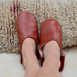 Peut inclure: Une paire de pantoufles en cuir brun rouge&acirc;tre. Les pantoufles sont ouvertes &agrave; l'arri&egrave;re et ont un bout arrondi. Les pantoufles sont sur une surface textur&eacute;e blanche et beige.
