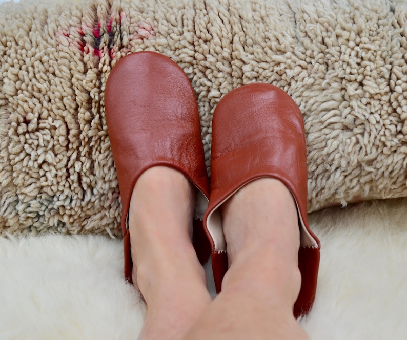 Peut inclure: Une paire de pantoufles en cuir brun rouge&acirc;tre. Les pantoufles sont ouvertes &agrave; l'arri&egrave;re et ont un bout arrondi. Les pantoufles sont sur une surface textur&eacute;e blanche et beige.