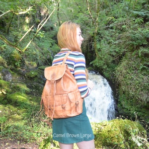 May include: A woman wearing a striped long-sleeved shirt and a green skirt is standing in a forest. She is wearing a large, brown leather backpack with multiple pockets and buckles. The backpack has the text "Camel Brown Large" printed on the bottom.