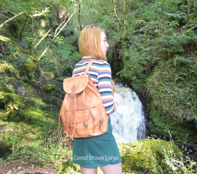 May include: A woman wearing a striped long-sleeved shirt and a green skirt is standing in a forest. She is wearing a large, brown leather backpack with multiple pockets and buckles. The backpack has the text "Camel Brown Large" printed on the bottom.
