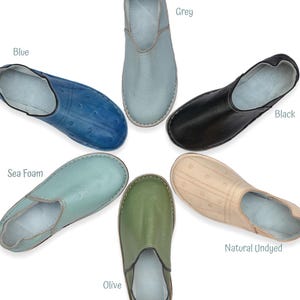 May include: Six slip-on shoes arranged in a circle, featuring colors labeled as Blue, Grey, Black, Natural Undyed, Olive, and Sea Foam. Each shoe has a smooth, rounded toe, a low-cut opening, and a subtle dotted pattern across the upper surface.