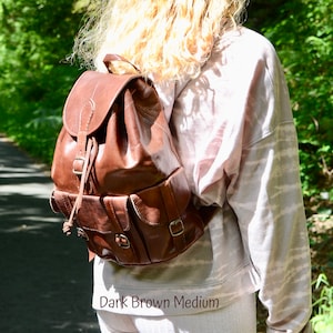 May include: A dark brown leather backpack with a drawstring closure and multiple pockets. The backpack is worn by a person with long blonde hair. The text "Dark Brown Medium" is visible at the bottom of the image.