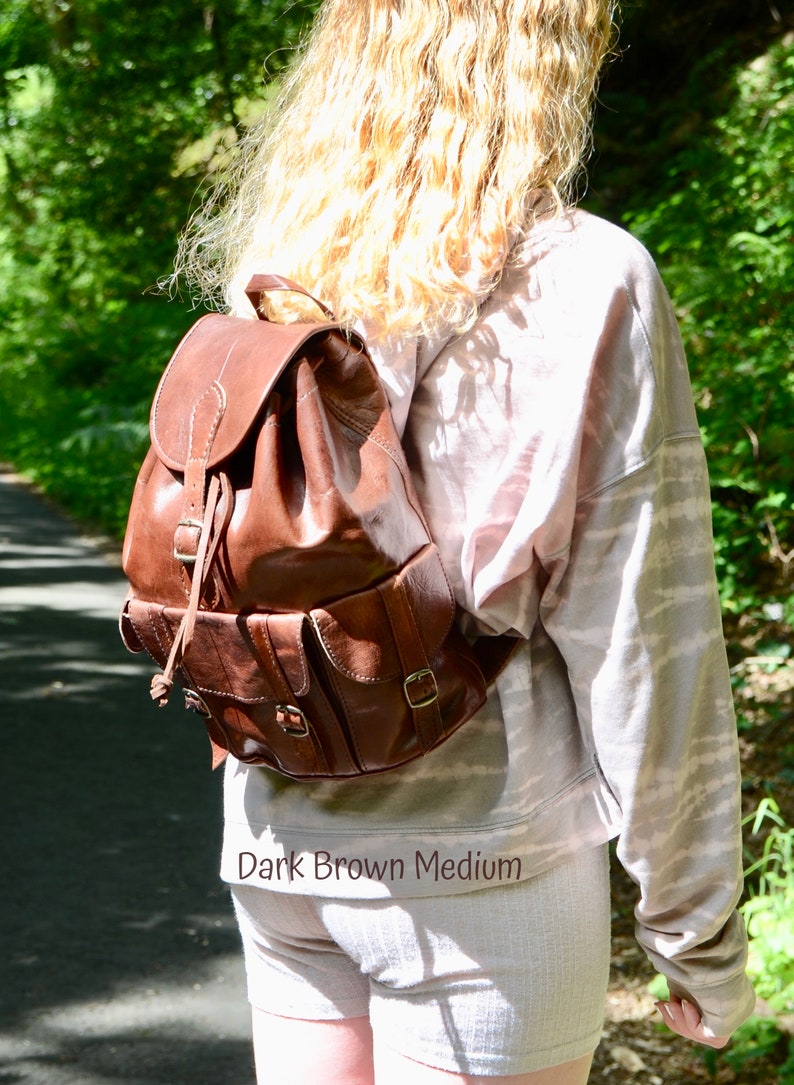 May include: A dark brown leather backpack with a drawstring closure and multiple pockets. The backpack is worn by a person with long blonde hair. The text "Dark Brown Medium" is visible at the bottom of the image.