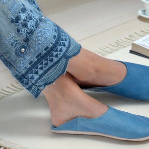 Peut inclure: Pantoufles en cuir bleu clair. Les pantoufles sont de couleur unie et ont un bout arrondi. L'image montre &eacute;galement un jean bleu avec des d&eacute;tails brod&eacute;s et un tapis blanc.