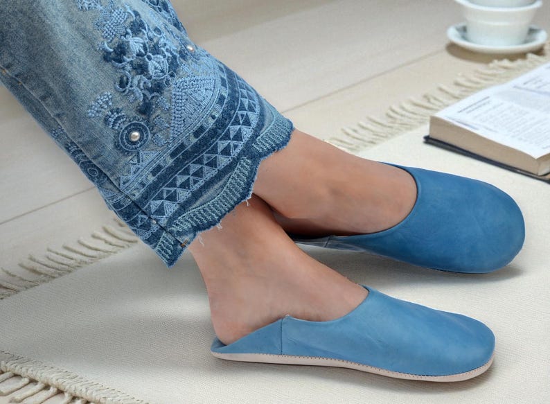 Peut inclure: Pantoufles en cuir bleu clair. Les pantoufles sont de couleur unie et ont un bout arrondi. L'image montre &eacute;galement un jean bleu avec des d&eacute;tails brod&eacute;s et un tapis blanc.