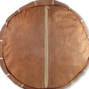 May include: A brown leather pouf with white stitching and a zipper closure. The pouf is round and has a smooth surface.