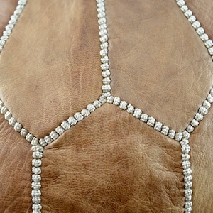 May include: Close-up of a brown leather pouf with white stitching. The stitching forms a geometric pattern.