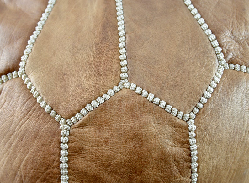 May include: Close-up of a brown leather pouf with white stitching. The stitching forms a geometric pattern.