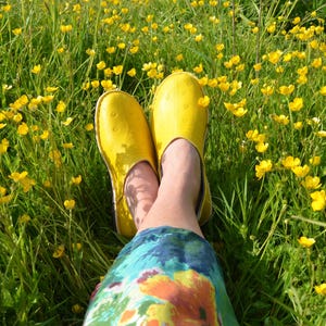 May include: A pair of bright yellow slip-on shoes with a smooth, rounded toe and a subtle dotted texture pattern. The shoes are worn on feet, which are positioned amidst a field of green grass and small yellow buttercup flowers in full bloom.