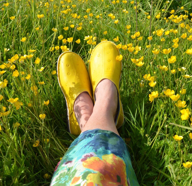 May include: A pair of bright yellow slip-on shoes with a smooth, rounded toe and a subtle dotted texture pattern. The shoes are worn on feet, which are positioned amidst a field of green grass and small yellow buttercup flowers in full bloom.