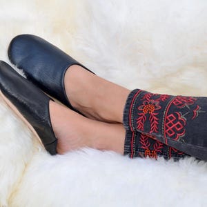 Peut inclure: Mocassins en cuir noir &agrave; bout arrondi. Les chaussons sont pos&eacute;s sur une surface blanche et duveteuse. La personne porte un jean en denim fonc&eacute; avec des motifs floraux brod&eacute;s rouges et oranges sur les poignets.