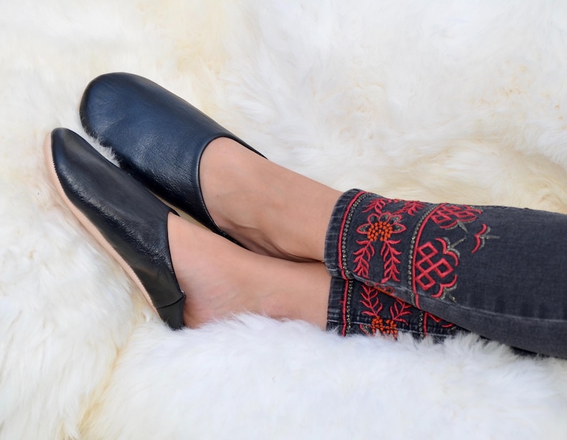 Peut inclure: Mocassins en cuir noir &agrave; bout arrondi. Les chaussons sont pos&eacute;s sur une surface blanche et duveteuse. La personne porte un jean en denim fonc&eacute; avec des motifs floraux brod&eacute;s rouges et oranges sur les poignets.