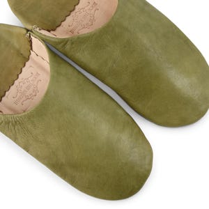 May include: A pair of olive green leather slippers with a rounded toe and a scalloped edge at the opening. The interior of each slipper features a stamped logo that reads ROCK THE KASBAH with decorative flourishes.
