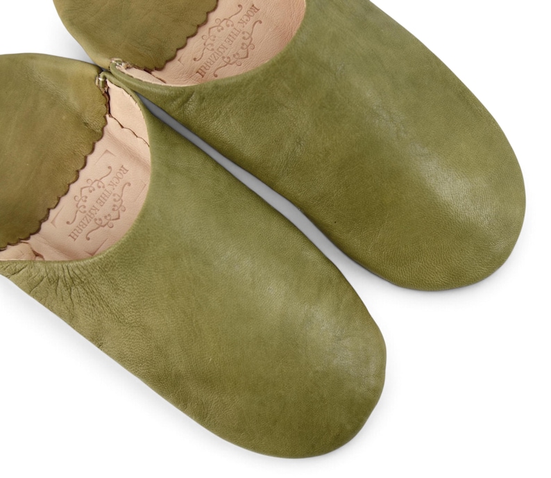 May include: A pair of olive green leather slippers with a rounded toe and a scalloped edge at the opening. The interior of each slipper features a stamped logo that reads ROCK THE KASBAH with decorative flourishes.