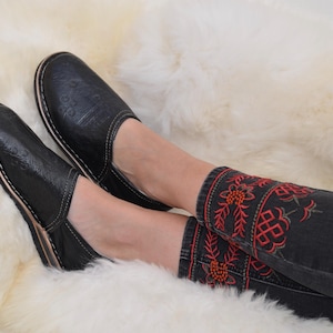 May include: A pair of black slip-on shoes with a subtle embossed pattern and white stitching along the sole, paired with dark gray jeans featuring red and green floral embroidery around the ankles, showcasing a detailed and textured design.