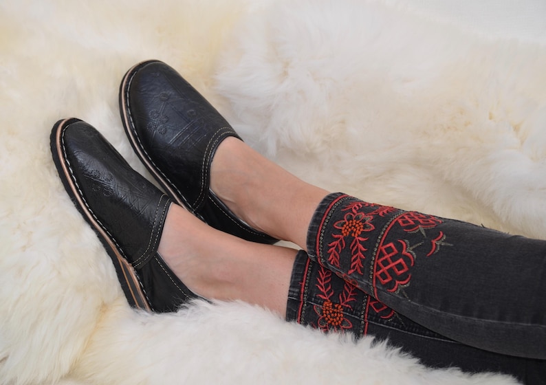 May include: A pair of black slip-on shoes with a subtle embossed pattern and white stitching along the sole, paired with dark gray jeans featuring red and green floral embroidery around the ankles, showcasing a detailed and textured design.