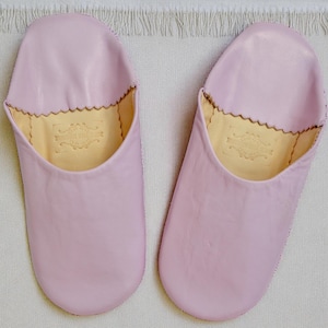 Peut inclure: Une paire de chaussons en cuir rose clair. Les chaussons ont un bout arrondi et un bord festonn&eacute; autour de l'ouverture. L'int&eacute;rieur est de couleur beige clair avec un embl&egrave;me d&eacute;coratif. Les chaussons sont sur une surface blanche textur&eacute;e.