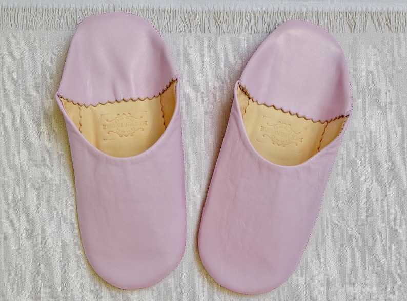 Peut inclure: Une paire de chaussons en cuir rose clair. Les chaussons ont un bout arrondi et un bord festonn&eacute; autour de l'ouverture. L'int&eacute;rieur est de couleur beige clair avec un embl&egrave;me d&eacute;coratif. Les chaussons sont sur une surface blanche textur&eacute;e.
