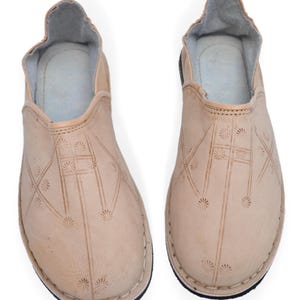 May include: A pair of light beige slip-on shoes featuring a rounded toe and a low-cut opening. The exterior surface is decorated with a repeating pattern of small, stamped sunburst motifs connected by thin, straight lines across the vamp.
