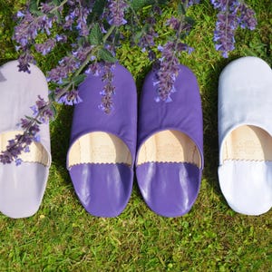 Peut inclure: Vue a&eacute;rienne de six pantoufles en cuir de couleurs lavande, violet et blanc, dispos&eacute;es sur de l'herbe verte. Les pantoufles ont un bout arrondi et un bord festonn&eacute;. Des fleurs violettes sont en arri&egrave;re-plan.