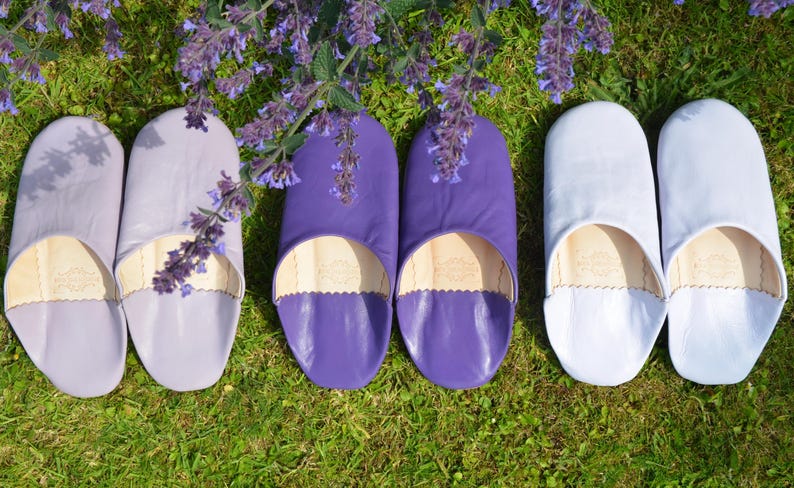 Peut inclure: Vue a&eacute;rienne de six pantoufles en cuir de couleurs lavande, violet et blanc, dispos&eacute;es sur de l'herbe verte. Les pantoufles ont un bout arrondi et un bord festonn&eacute;. Des fleurs violettes sont en arri&egrave;re-plan.