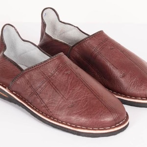 May include: A pair of dark brown leather slippers featuring a textured surface with subtle stitched cross patterns on the upper. The slippers have a high-cut heel collar, a light-colored interior lining, and a layered sole with a black tread.