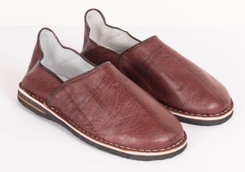 May include: A pair of dark brown leather slippers featuring a textured surface with subtle stitched cross patterns on the upper. The slippers have a high-cut heel collar, a light-colored interior lining, and a layered sole with a black tread.
