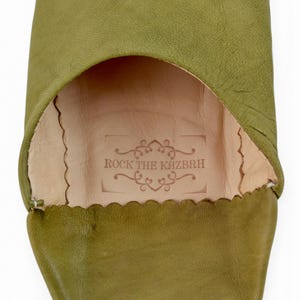 May include: An olive green leather slipper with a rounded toe and a scalloped edge opening. The interior features a light beige leather insole stamped with the text ROCK THE KAZBAH surrounded by decorative scrollwork and a rectangular border.