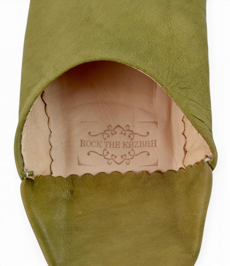 May include: An olive green leather slipper with a rounded toe and a scalloped edge opening. The interior features a light beige leather insole stamped with the text ROCK THE KAZBAH surrounded by decorative scrollwork and a rectangular border.