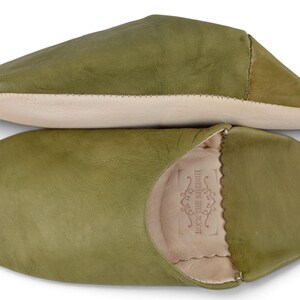 May include: A pair of olive green leather slippers with a smooth finish. One slipper is shown from the side, and the other is shown from the top, revealing a light beige interior lining and a stamped logo that reads ROCK THE KASBAH.