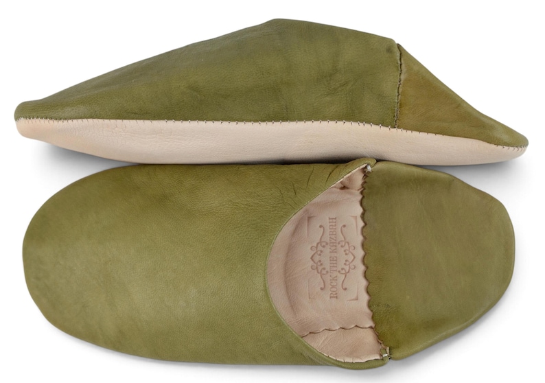 May include: A pair of olive green leather slippers with a smooth finish. One slipper is shown from the side, and the other is shown from the top, revealing a light beige interior lining and a stamped logo that reads ROCK THE KASBAH.