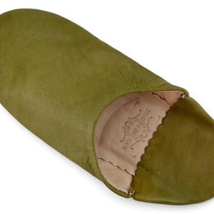 May include: An olive green leather slipper with a rounded toe and a soft, light beige interior lining. The inner sole features a stamped logo with the text ROCK THE KASBAH surrounded by decorative scrollwork and a scalloped edge.