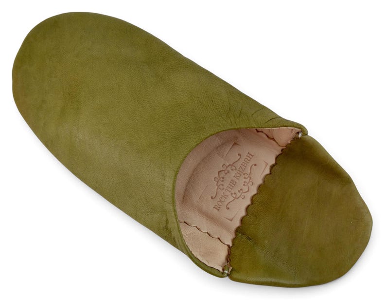 May include: An olive green leather slipper with a rounded toe and a soft, light beige interior lining. The inner sole features a stamped logo with the text ROCK THE KASBAH surrounded by decorative scrollwork and a scalloped edge.