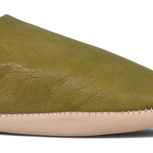 May include: A close-up view of an olive green leather slipper featuring a smooth, rounded toe and a light beige sole. The upper portion of the slipper is crafted from a single piece of textured leather, joined to the sole with visible stitching.