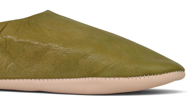 May include: A close-up view of an olive green leather slipper featuring a smooth, rounded toe and a light beige sole. The upper portion of the slipper is crafted from a single piece of textured leather, joined to the sole with visible stitching.