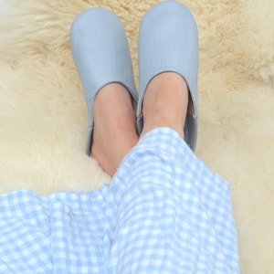 Peut inclure: Une paire de pantoufles en cuir bleu clair. Les pantoufles sont pos&eacute;es sur une surface moelleuse de couleur cr&egrave;me. La personne porte un pyjama &agrave; carreaux bleu clair et blanc.