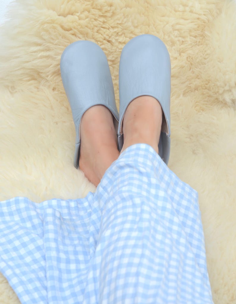 Peut inclure: Une paire de pantoufles en cuir bleu clair. Les pantoufles sont pos&eacute;es sur une surface moelleuse de couleur cr&egrave;me. La personne porte un pyjama &agrave; carreaux bleu clair et blanc.