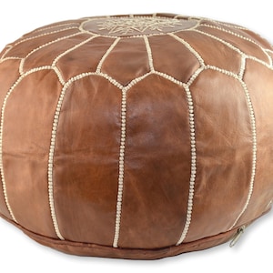 May include: A brown leather pouf with white stitching and a decorative design on the top. The pouf has a zipper on the side.