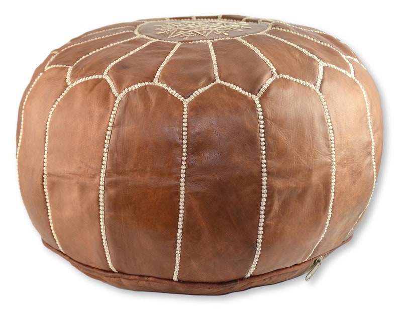 May include: A brown leather pouf with white stitching and a decorative design on the top. The pouf has a zipper on the side.
