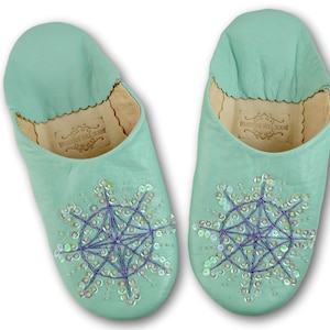 May include: A pair of mint green leather slippers with a snowflake design embroidered with iridescent sequins and purple thread. The slippers have a leather insole and a label that reads "Rock the Kasbah".