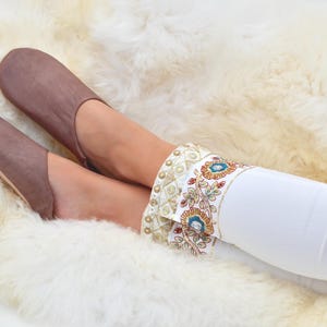Peut inclure: Des chaussons en cuir marron &agrave; bout pointu, port&eacute;s avec un pantalon blanc orn&eacute; d'un bracelet d&eacute;coratif avec des accents floraux et dor&eacute;s. Les chaussons reposent sur un tapis blanc et moelleux. Les chaussons sont en cuir.