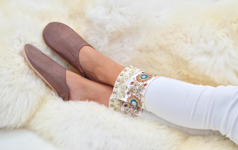 Peut inclure: Des chaussons en cuir marron &agrave; bout pointu, port&eacute;s avec un pantalon blanc orn&eacute; d'un bracelet d&eacute;coratif avec des accents floraux et dor&eacute;s. Les chaussons reposent sur un tapis blanc et moelleux. Les chaussons sont en cuir.