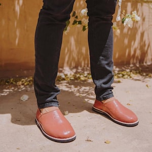 May include: A pair of slip-on shoes in a muted terracotta color with a smooth, rounded toe design and a thin, dark-colored sole. The shoes feature a low-cut opening and a simple, unadorned exterior surface, worn with dark-colored denim jeans.